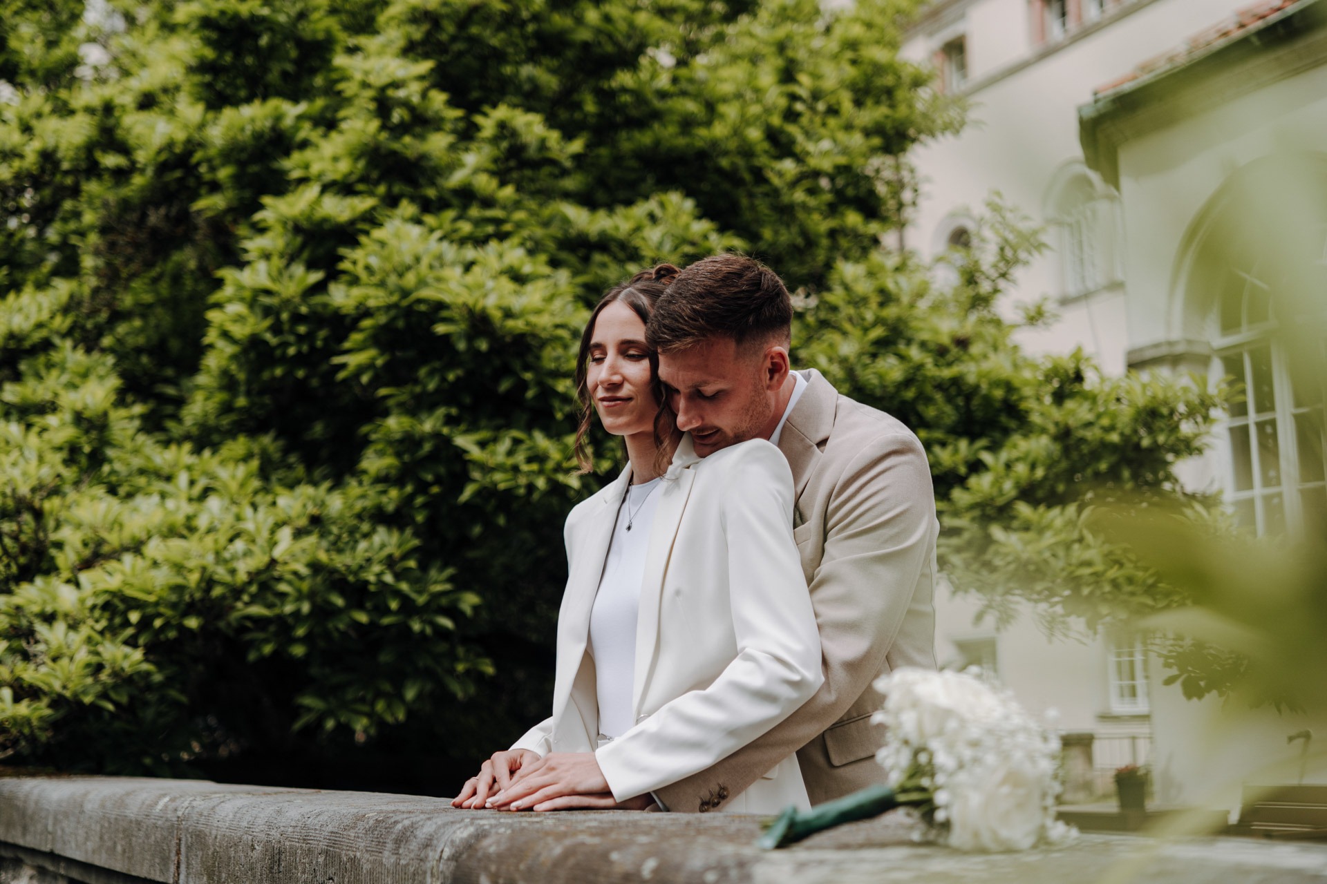 Melissa und Lukas stehen gemeinsam an einer Steinbrüstung vor sommerlichem Grün.