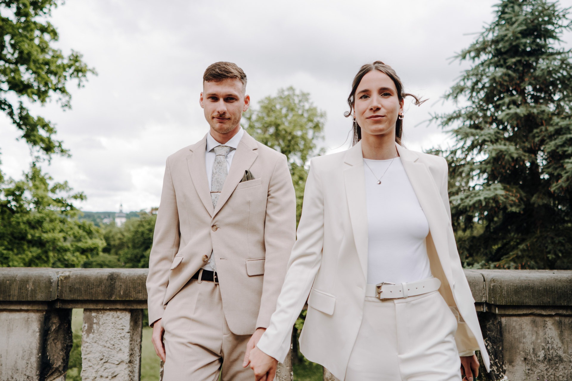 Melissa und Lukas gehen Hand in Hand entlang einer steinernen Balustrade im Grünen.