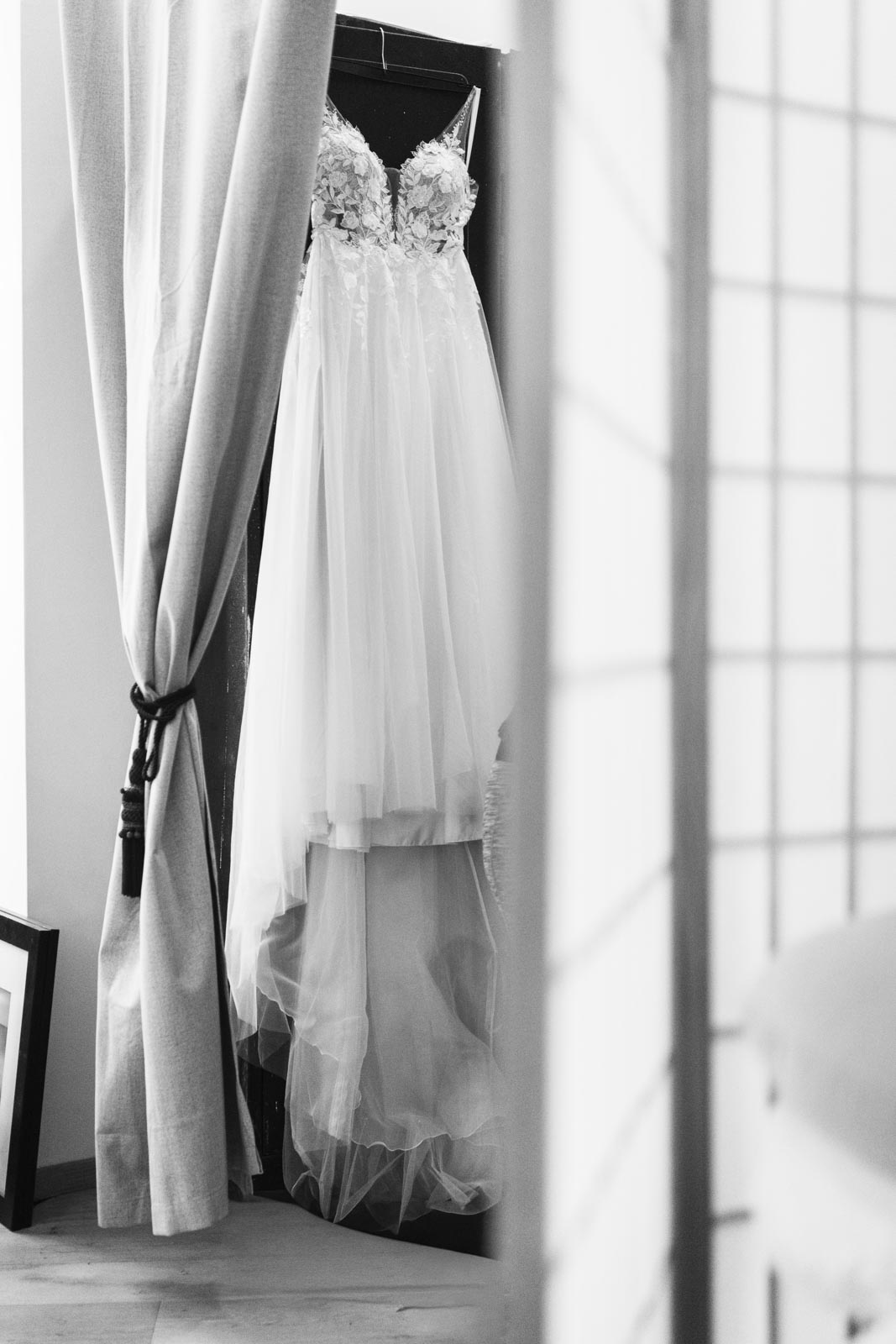 Schwarz-Weiß-Aufnahme eines Brautkleides, das vor einem hellen Fenster zwischen Vorhängen hängt.