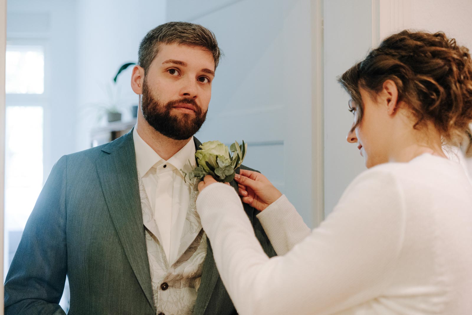 Luisa befestigt eine helle Ansteckblume an Jonas' grünem Sakko.