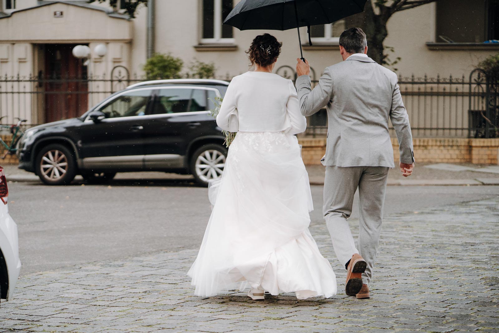 Luisa geht mit einem Begleiter im hellen Anzug durch den Regen über die Straßenn Leipzig.
