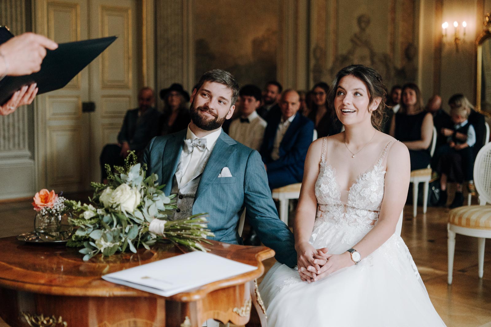 Luisa und Jonas sitzen nebeneinander an einem kleinen Tisch im historischen Trausaal und blicken nach vorn.