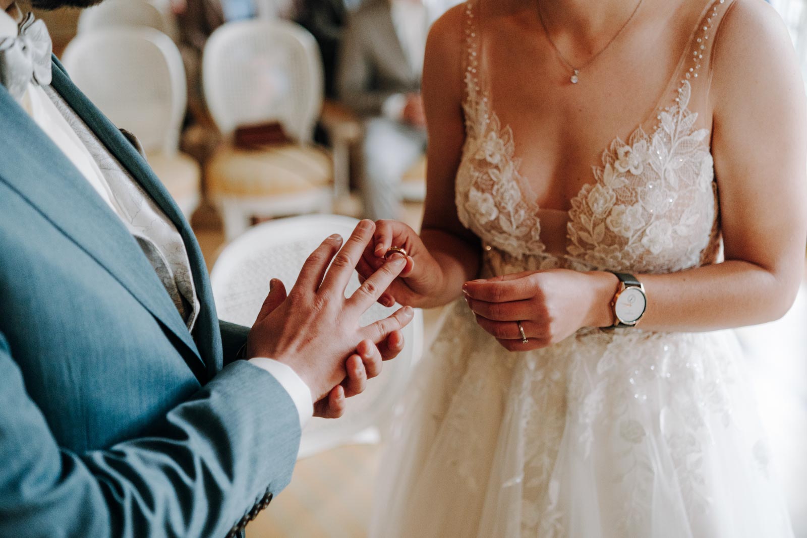 Nahaufnahme der Hände von Luisa und Jonas beim Ringwechsel, mit Brautkleid und grünem Anzug im Ausschnitt.