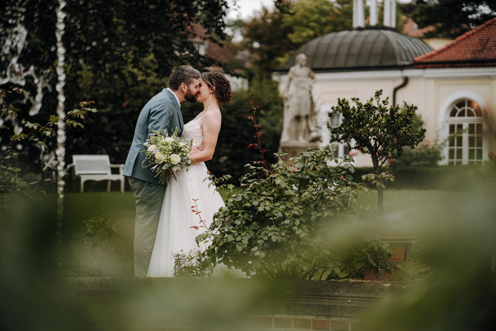Luisa und Jonas küssen sich im Schlossgarten, grüne Blätter liegen unscharf im Vordergrund.