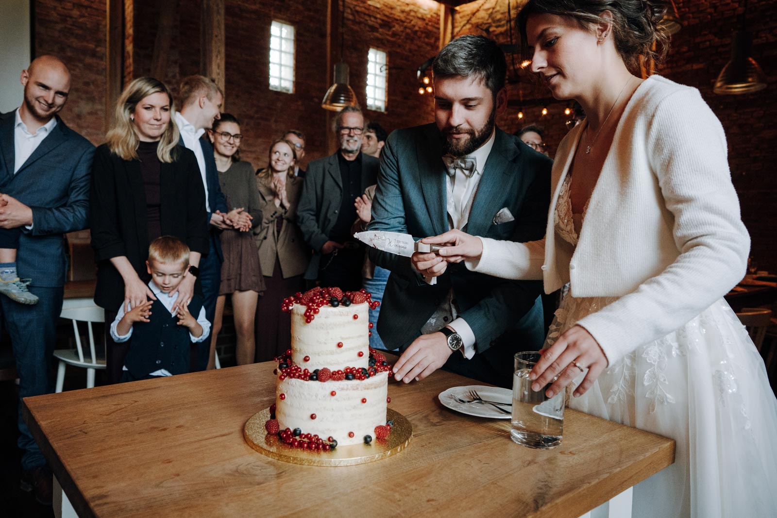 Luisa und Jonas schneiden gemeinsam ihre zweistöckige Hochzeitstorte an, umringt von Gästen.