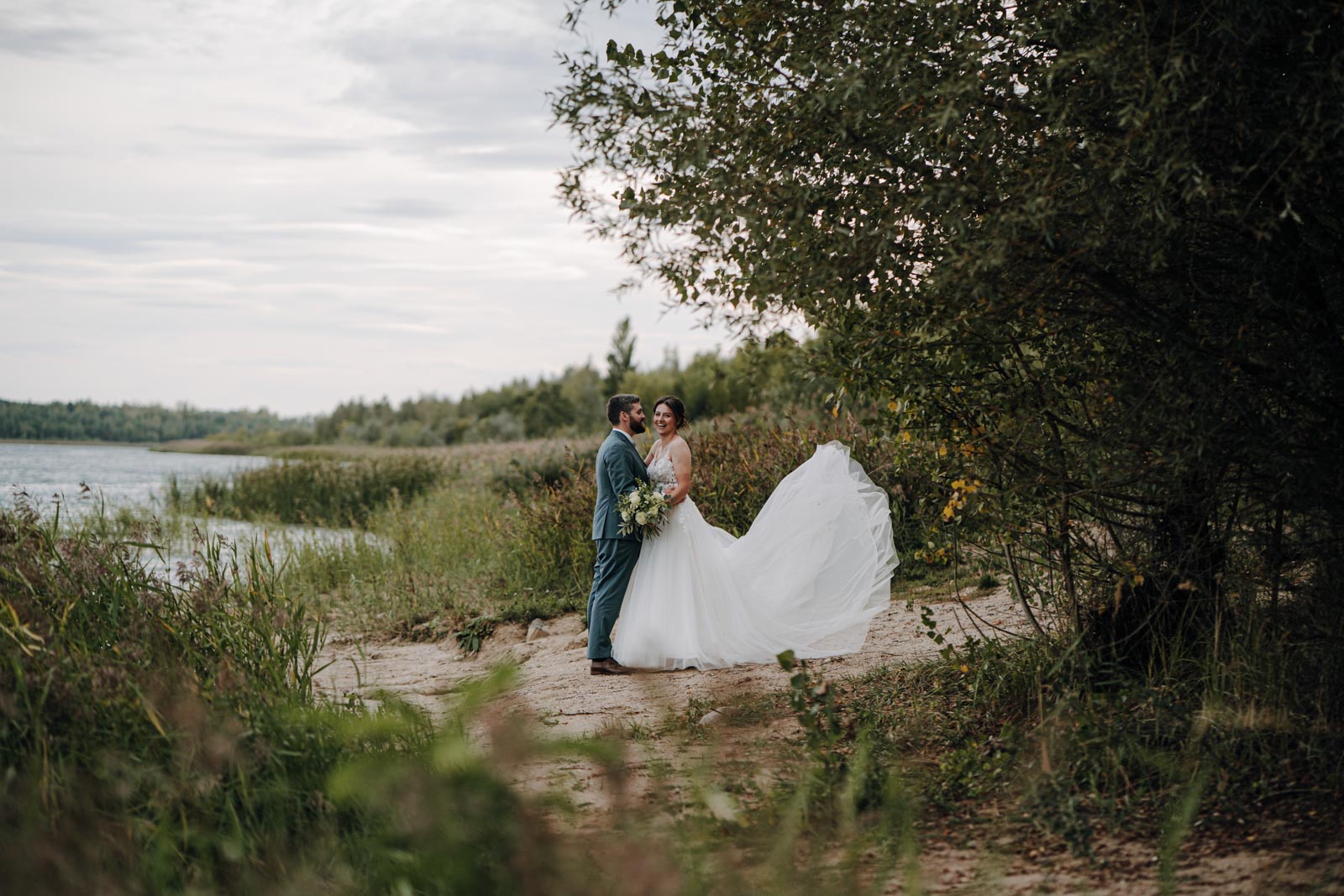 Luisa und Jonas stehen am Ufer zwischen Bäumen und Schilf, Luisas Brautkleid bewegt sich seitlich.