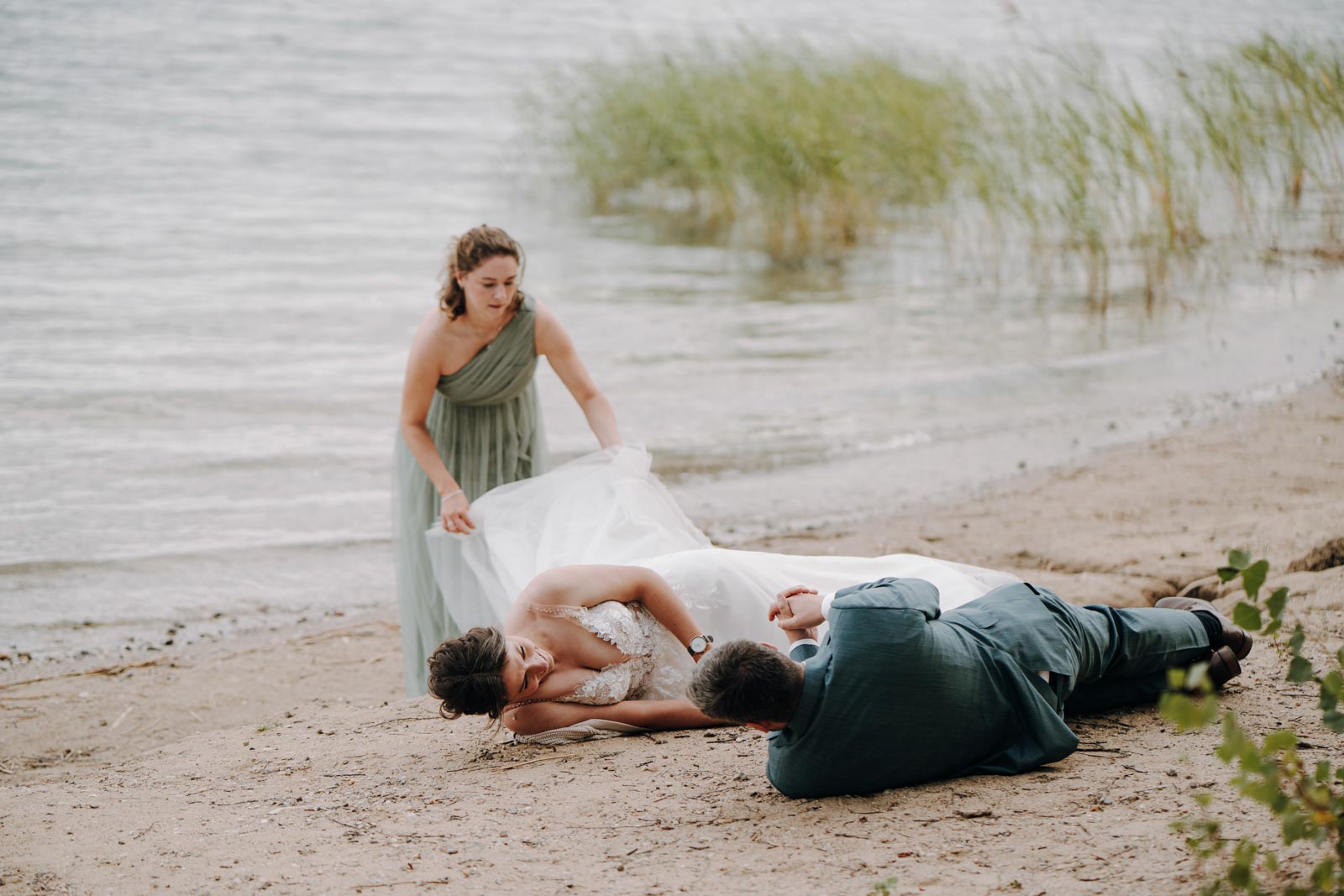 Luisa und Jonas liegen nebeneinander im Sand am Wasser, eine Begleiterin steht hinter ihnen.