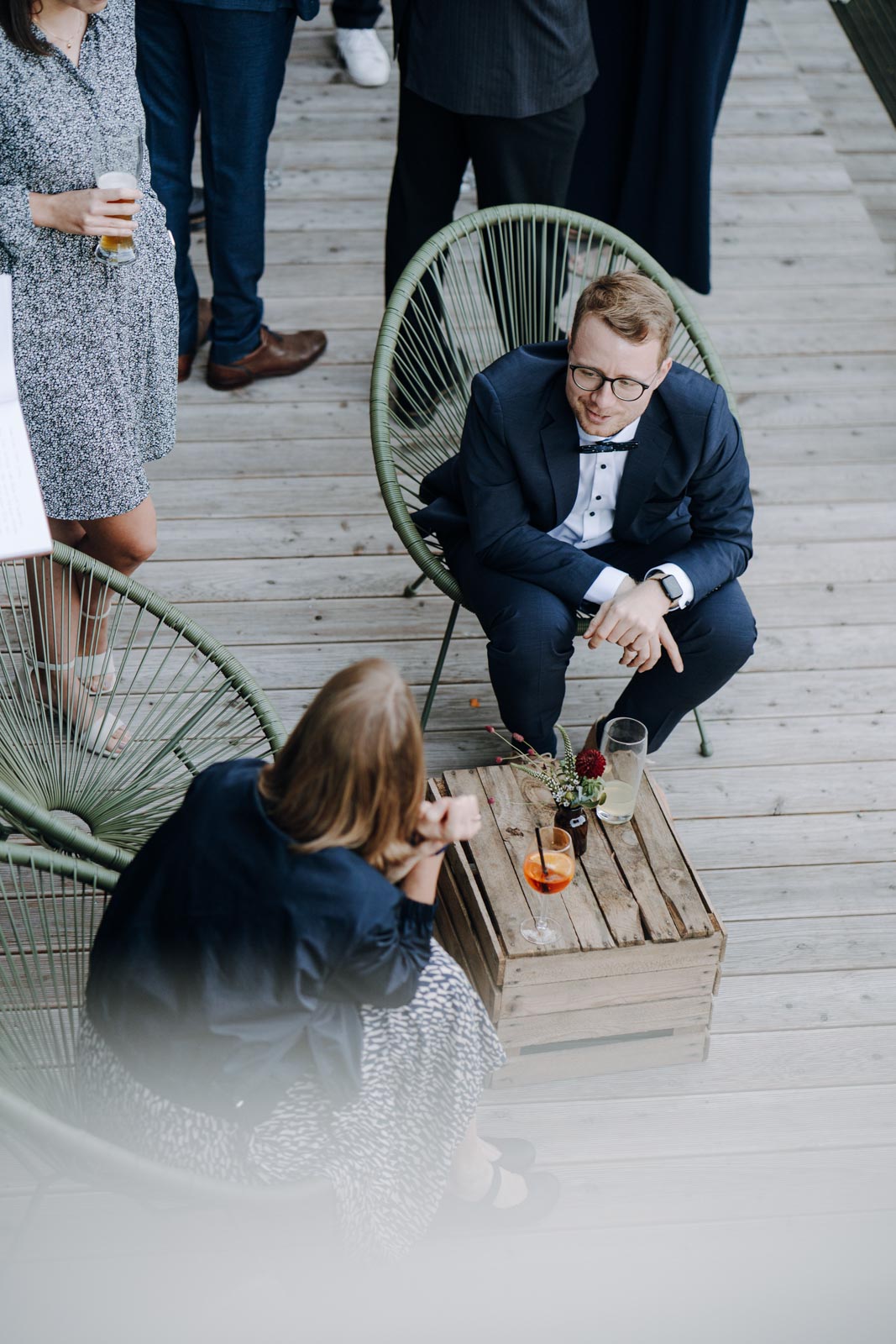 Hochzeitsgäste sitzen mit Getränken an einem kleinen Tisch im Freien, aus erhöhter Perspektive fotografiert.
