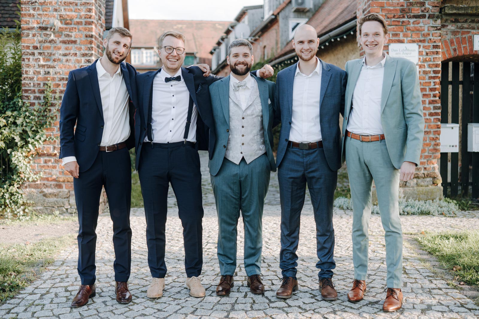 Jonas steht mit vier Freunden in Anzügen vor einem Backsteingebäude und legt die Arme um ihre Schultern.
